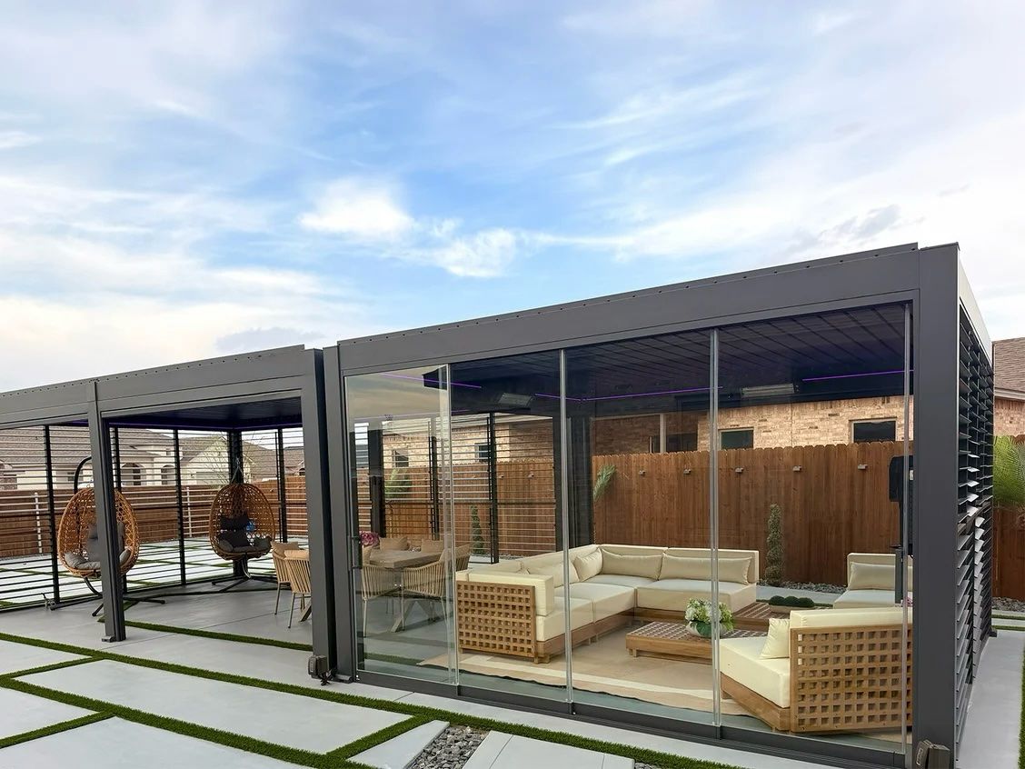 Two joined pergolas side by side, one enclosed with glass walls around a cream sectional sofa, the other open with hanging egg chairs